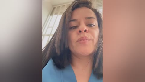 Video of a woman with medium skin tone and shoulder-length dark hair, wearing a light blue top. Background features a blurred, bright room with large windows.