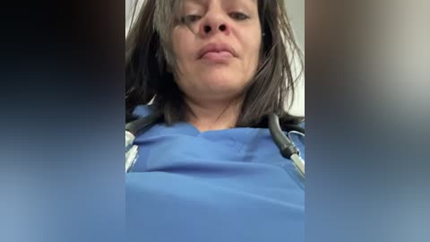 Video of a woman with shoulder-length brown hair, wearing a blue surgical gown, with a stethoscope around her neck, taken from a low angle.