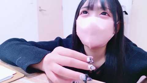 Video of an Asian woman with straight black hair, wearing a black mask, a black sweater, and white nail polish. She is seated indoors, leaning forward, looking directly at the camera.