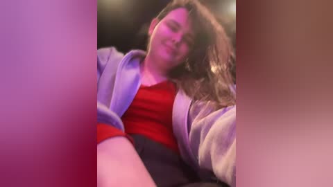 A video of a young woman with long brown hair, wearing a red top and grey hoodie, lying on her side, slightly blurred and out of focus.