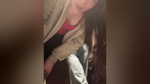 A blurry video shows a person with long brown hair, wearing a beige cardigan over a red shirt, leaning over, possibly towards a person in gray pants.