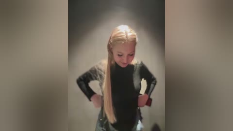 Video of a blonde woman with long hair in a black, textured jacket, hands on hips, standing in a dimly lit, narrow hallway.