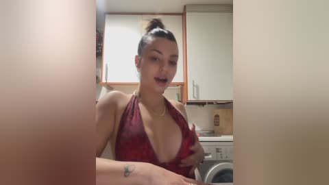 Video of a curvy, light-skinned woman with dark hair tied in a bun, wearing a revealing red halter top, sitting in a modern, beige kitchen.