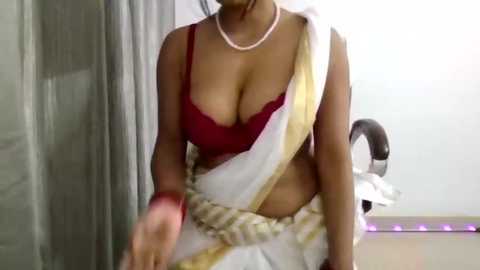 Video of a woman in a red bra and white sari with gold border, wearing a pearl necklace, indoors.