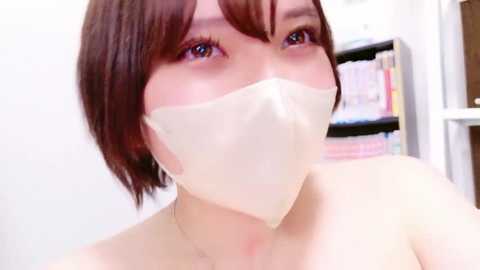 A video of a young East Asian woman with short, brown hair, wearing a white face mask, in a white room with bookshelves and colorful books in the background.