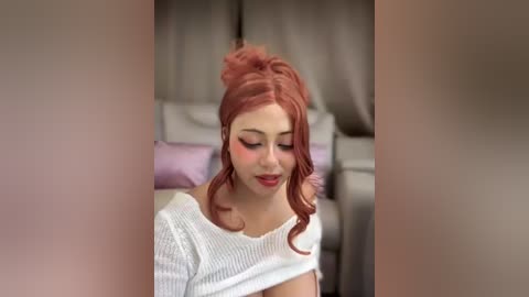 Video of a young woman with fair skin and long, wavy, auburn hair styled in a high bun. She wears a white, ribbed knit top and sits indoors on a gray leather couch, surrounded by light-colored cushions.