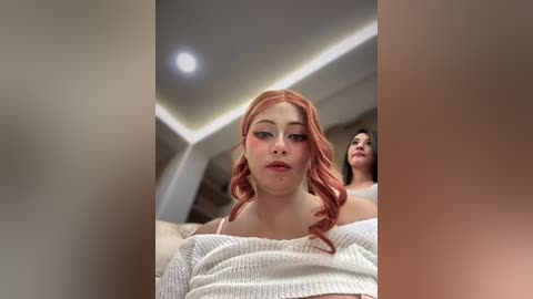 Video of a young woman with pale skin and long, wavy, pink hair, wearing a white, off-shoulder sweater. She stands in a modern, dimly-lit room with recessed lighting, blurred background.