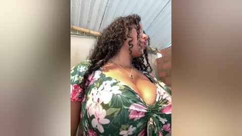 A video of a plus-size Black woman with curly hair and a large chest, wearing a low-cut floral dress, standing indoors under a corrugated metal roof.