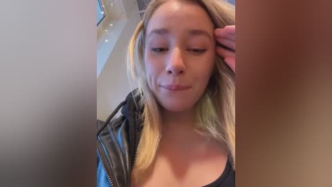 Video of a young woman with light skin and straight blonde hair, wearing a black leather jacket and a black top, touching her hair, indoors with a blurred background.