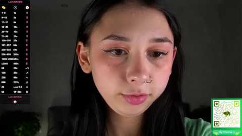Video of a young Asian woman with long black hair, light skin, and a silver nose ring, wearing a green top, in a dimly lit room.