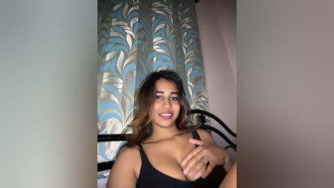 Video of a young woman with medium brown skin, wearing a black tank top, smiling and covering her breast. Floral-patterned curtains in the background.