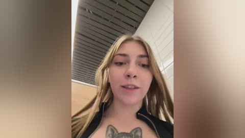 A young woman with long blonde hair, fair skin, and a black jacket, stands in a dimly lit, modern hallway with a dark ceiling and white tiled walls. She has a gray wolf tattoo on her chest.