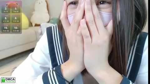Video of a young Asian woman with straight, dark hair, wearing a white sailor uniform, face mask, and hands clasped in prayer, indoors.