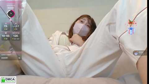 A video showing a young woman with a face mask, wearing white pajamas, lying on a bed, with a smartphone displaying a time of 10:50.