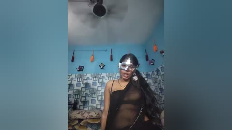 Video of a dark-skinned woman with long black hair in a black sheer dress, wearing a reflective mask and necklace, standing in a blue-walled room with patterned wallpaper.