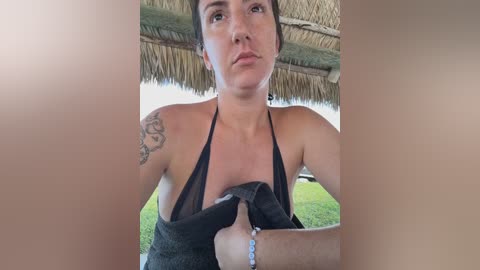 Video of a light-skinned woman with short brown hair, wearing a black halter top, with a tattoo on her left arm, standing in a thatched-roof hut.