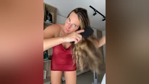 Video of a blonde woman with fair skin, wearing a red tank top and no pants, holding a hairbrush, in a modern bathroom with beige walls and shelves.