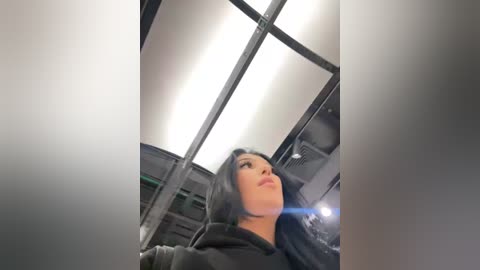 A video of a woman with long, black hair, wearing a dark hoodie, standing inside a brightly lit, modern, glass-walled elevator with fluorescent lighting.