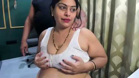 Video of a plus-size South Asian woman with fair skin, wearing a white tank top, holding her large breasts. She has a relaxed, confident expression. Background features a green wall, a man in a black shirt, and patterned curtains.
