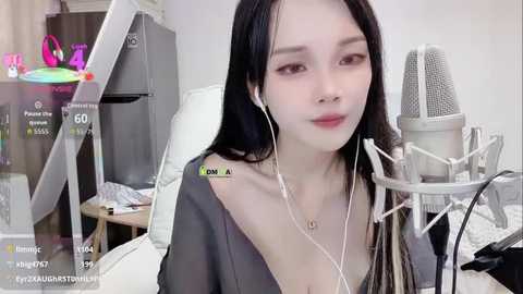 Video of a young East Asian woman with long black hair, wearing headphones, sitting in front of a microphone. Background includes a colorful toy, a desk, and a chair.