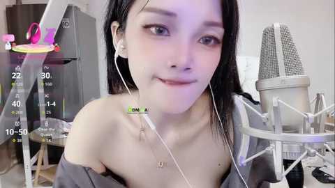 Video of an East Asian woman with fair skin, straight black hair, wearing a gray off-shoulder top, sitting in front of a microphone, holding a green light stick, in a home studio with a grey fridge and white wall.