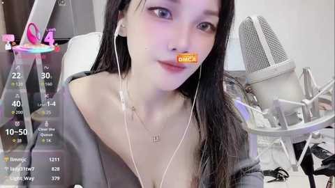 Video of a young Asian woman with long black hair, wearing a grey off-shoulder top, headphones, and a microphone, sitting at a cluttered desk with a white chair, computer, and a cat bed.