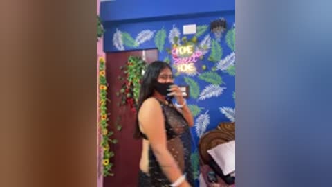 Video of a woman with medium skin tone, long black hair, wearing a black lace dress and face mask, taking a mirror selfie in a colorful room with blue walls, green leaf art, and a sunflower garland.