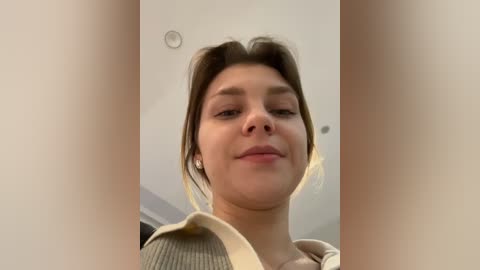Video of a young Caucasian woman with fair skin and brown hair, wearing a beige shirt, indoors with a white ceiling.