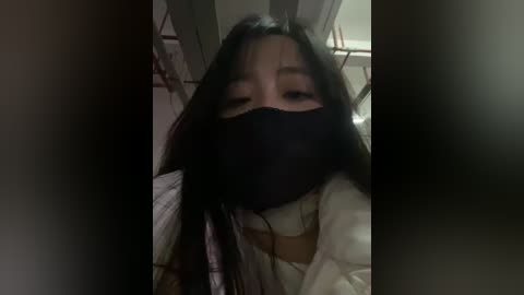 Video of a young Asian woman with long black hair, wearing a black face mask, white shirt, and dark jacket. The dimly lit room has a metal railing in the background.