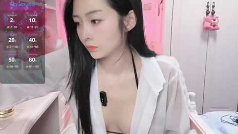 Video of an Asian woman with long black hair, wearing a white lab coat, sitting at a pink desk in a room with a digital display showing temperature and humidity.