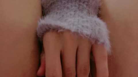 A close-up video of a hand with light skin, wearing a fluffy, grayish-purple sleeve, gently pressing against a light beige wall. The texture of the sleeve appears soft and cozy, contrasting with the smooth surface of the wall.
