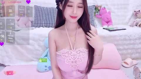 Video of a young East Asian woman with long black hair and fair skin, wearing a pastel pink lace dress, kneeling in a pastel-themed room with a plush toy, cushions, and a bed in the background.