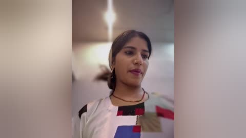 Video of a young woman with medium skin tone, dark hair, wearing a colorful patchwork blouse, standing in a dimly lit corridor with soft lighting.