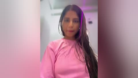 Video of a young woman with long, wet black hair, wearing a pink shirt, standing in a dimly lit bathroom with purple lighting.