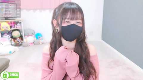 Video of an Asian woman with long black hair, wearing a black face mask and a pink off-shoulder top, kneeling in a soft pink room with plush toys and a TV in the background.