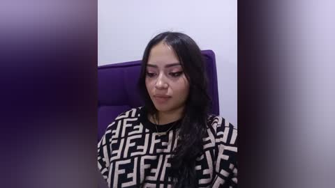 Video of a young woman with long, dark hair, wearing a black and white patterned top, seated in a purple chair, with a blurred background.