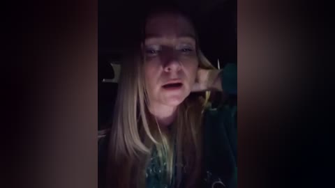 A dimly lit video of a middle-aged woman with fair skin and long blonde hair, wearing a green sweater, looking distressed and speaking into a phone.