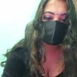 A close-up video of a woman with long, curly brown hair, wearing a black mask, and a black top. Her face is partially obscured, with a hint of pink lipstick visible. The background is a soft gradient of light blue and white.