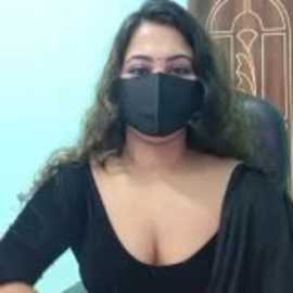 Video of a young woman with long brown hair, wearing a black face mask and black top, sitting in a room with wooden doors and light blue walls.