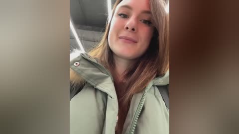 A video of a young Caucasian woman with straight, light brown hair, wearing a green winter jacket, smiling. The background shows the car interior with a blurry, neutral-toned headrest.