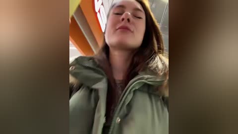 A video of a woman with light skin and long brown hair wearing a green coat, standing indoors with closed eyes and a relaxed expression.