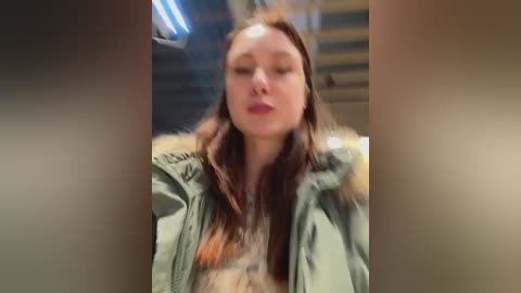 A blurry video of a woman with fair skin, long brown hair, and closed eyes, wearing a green jacket, in a dimly lit room with metallic surfaces.
