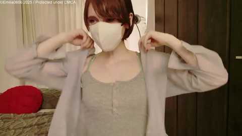 Video of a pale, slender woman with short auburn hair, wearing a face mask, a light green tank top, and a white cardigan. She adjusts the cardigan's sleeves. Background includes a bed with red and beige blankets, and wooden panels.