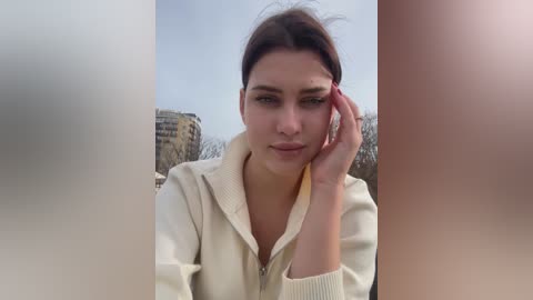 Video of a woman with fair skin, brown hair, and blue eyes, wearing a beige sweater, resting her chin on her hand, against a blurred urban background.