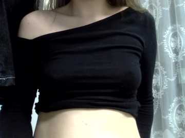 Video of a light-skinned woman with long, straight brown hair, wearing a black off-the-shoulder crop top that accentuates her medium-sized breasts. She stands in a bathroom with light-colored, patterned curtains in the background.