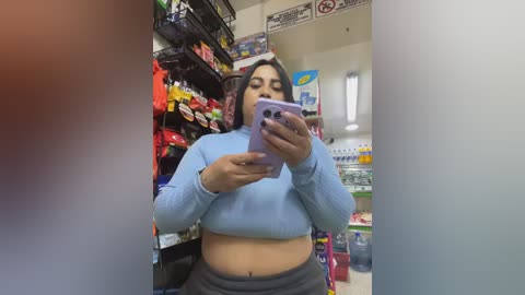 A video of a Latina woman with medium skin tone, medium build, and shoulder-length dark hair in a grocery store. She wears a light blue crop top and gray pants, holding a lavender phone, capturing a mirror selfie.