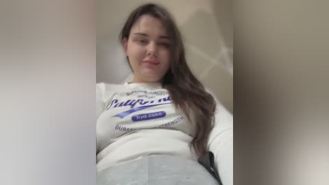 Video of a young woman with long brown hair, fair skin, wearing a white long-sleeve shirt with blue text and gray pants, standing indoors against a beige wall.