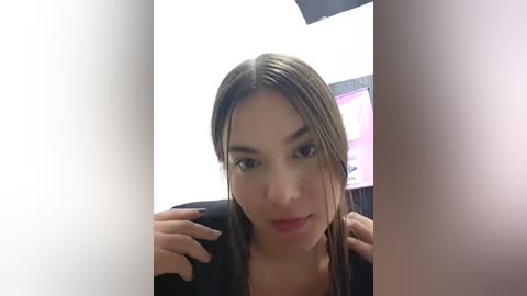 A young woman with light brown skin, brown hair, and a small nose, wearing a black top, sits in front of a computer screen with a blurred image.