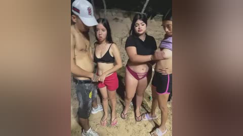 Video of four people standing on a sandy beach at night. Two women in sports bras and shorts, a man in a black shirt, and a woman in a purple bikini.