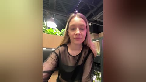 Video of a young Caucasian woman with straight, shoulder-length blonde hair, wearing a sheer black top, sitting indoors with green plants and industrial lighting in the background.